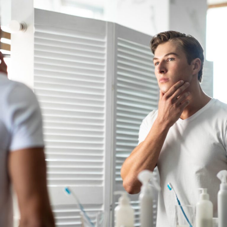 Serious handsome man touching his face, looking in the mirror in the bathroom